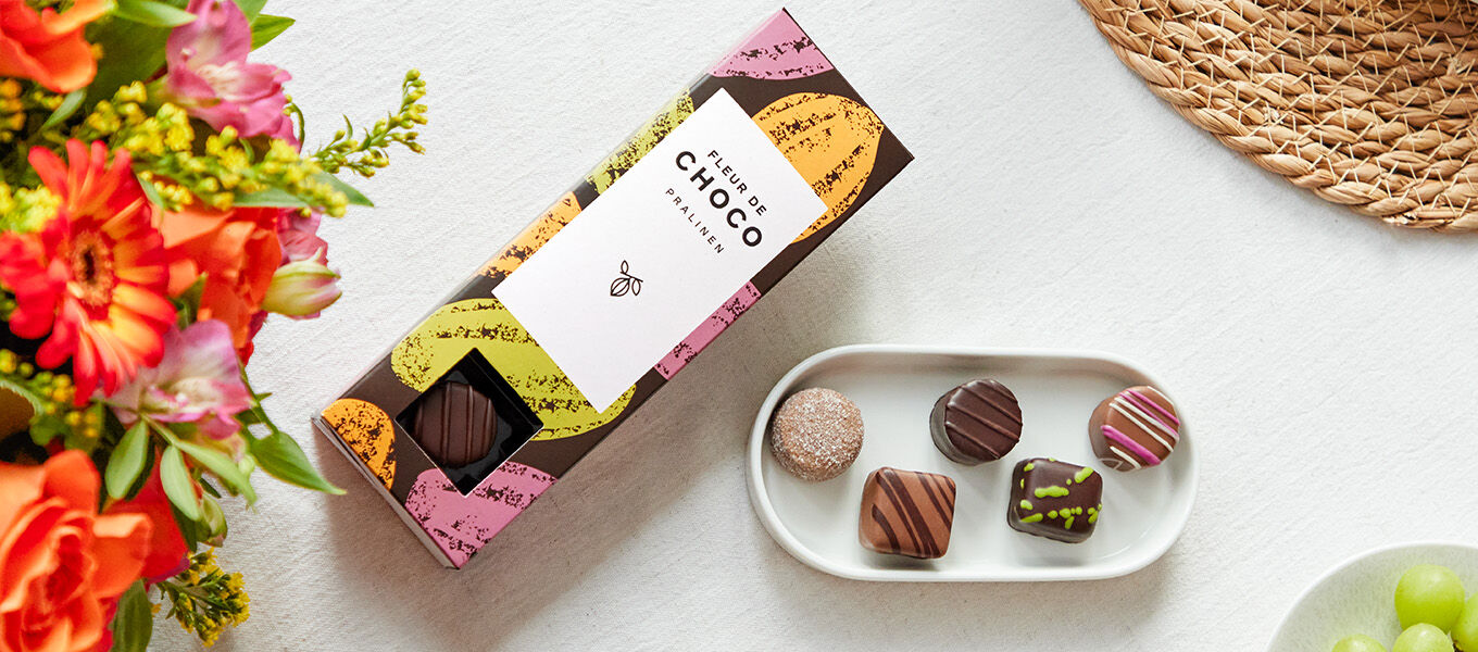 Offene Schachtel „Lindt Süße Momente“ Pralinen in edler schwarzer Verpackung mit goldenen Pflanzenmustern, daneben ein bunter Blumenstrauß mit orangefarbenen Rosen, gelben und violetten Blüten.